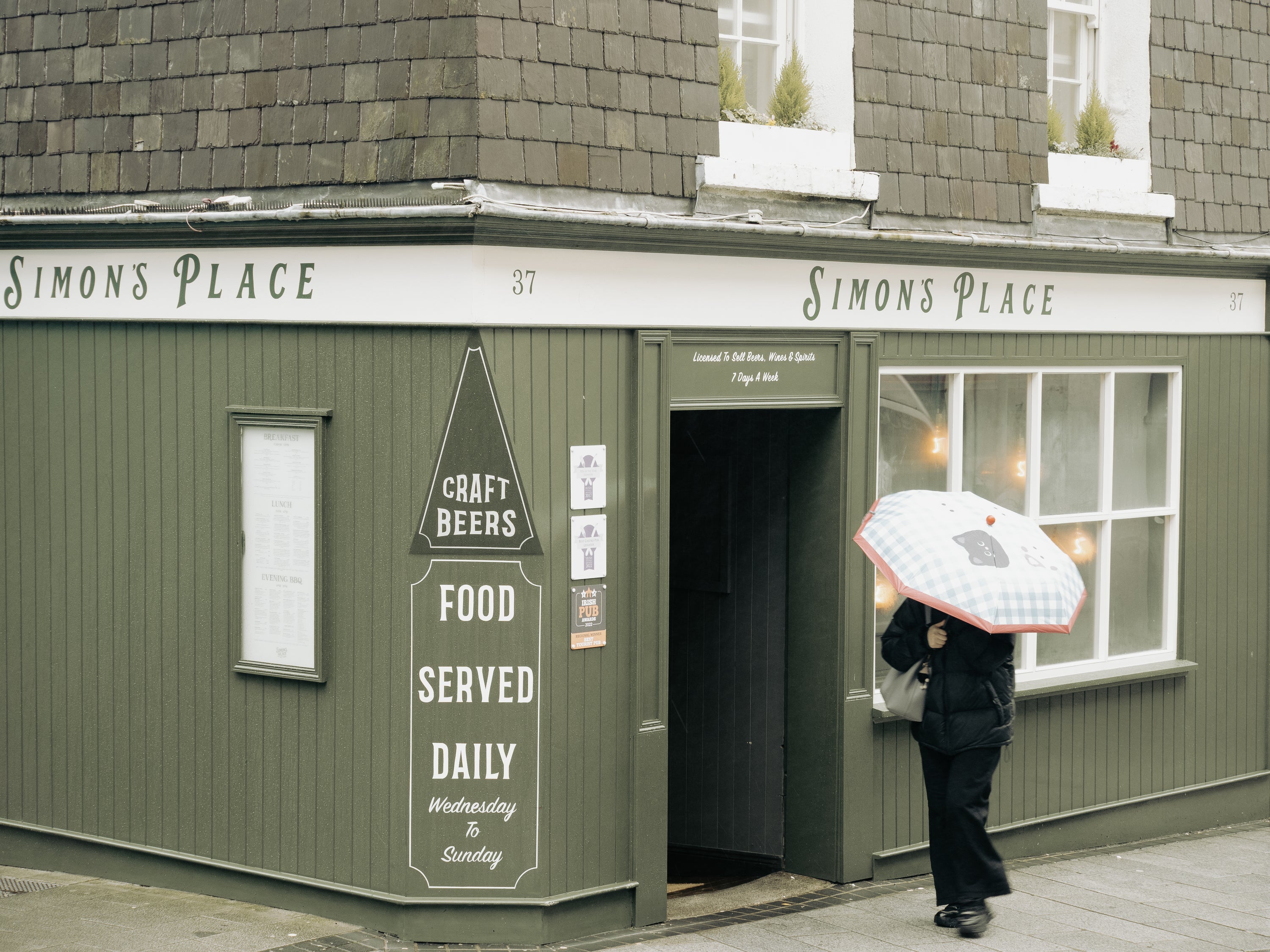 Person with an umbrella standing outside a building labeled 'Simon's Place' with a sign for craft beers and food.